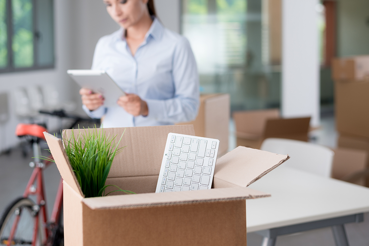 Box of office supplies on desk with business woman using tablet in background.
