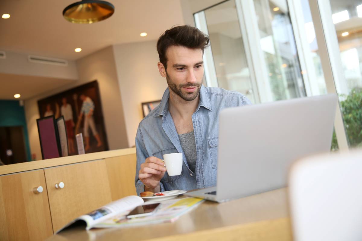 A man working remotely at a table drinking coffee and using a laptop